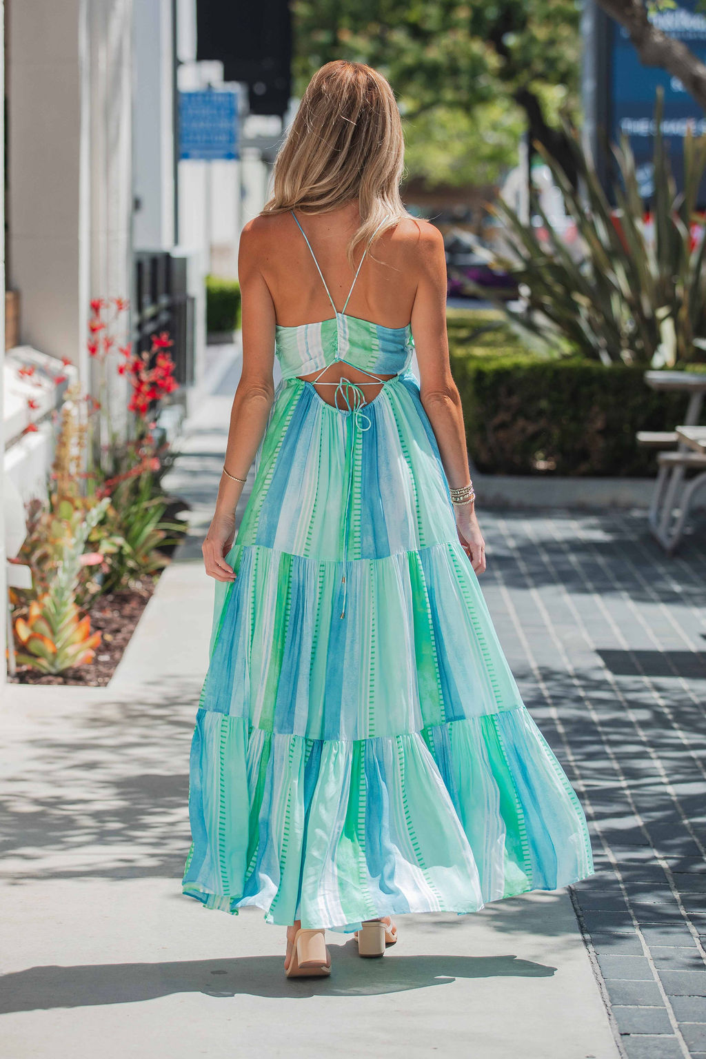 A woman in an ELAN Seafoam Print Tiered Maxi Dress with an adjustable back tie walks on a sunny sidewalk with plants and buildings.
