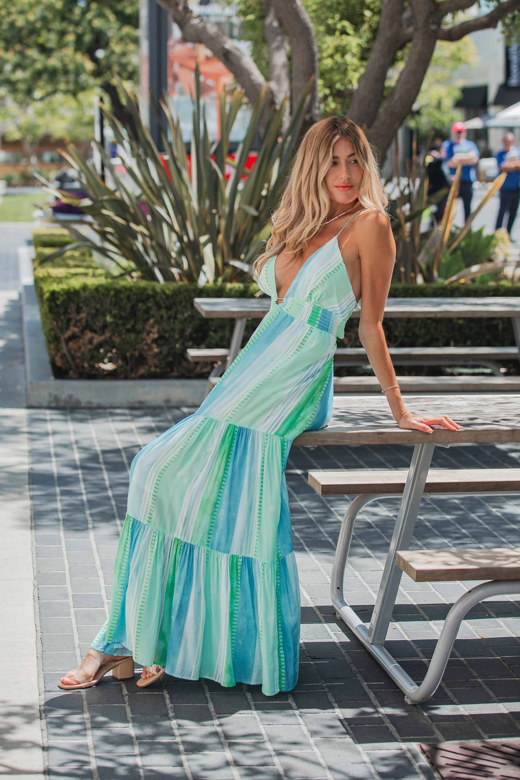 A woman in the ELAN Seafoam Print Tiered Maxi Dress with an adjustable back tie leans on a picnic table outdoors among lush greenery.