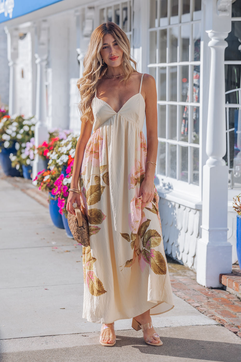 A woman in the Buttercup Floral Print Maxi Dress and sandals stands on a sidewalk, holding a brown clutch, with white buildings behind her.