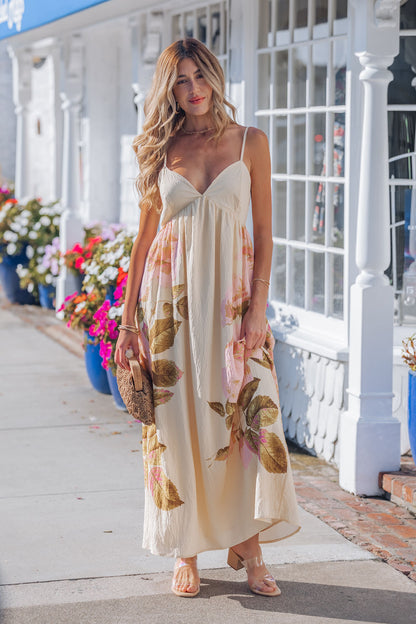 A woman in the Buttercup Floral Print Maxi Dress and sandals stands on a sidewalk, holding a brown clutch, with white buildings behind her.