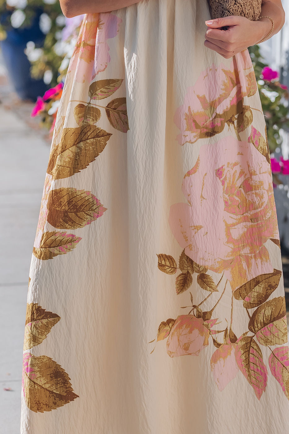 A woman in the Buttercup Floral Print Maxi Dress with an empire waist stands on a sunny sidewalk, effortlessly elegant.