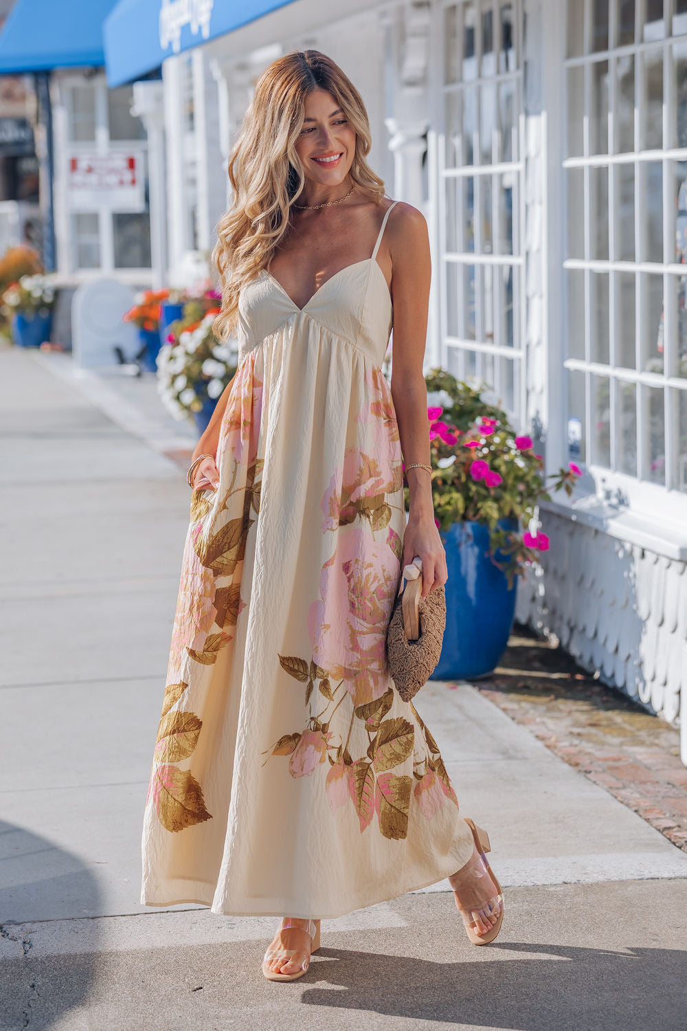 Woman smiles in the Buttercup Floral Print Maxi Dress with adjustable cami straps, walking past bright flowers and white buildings.