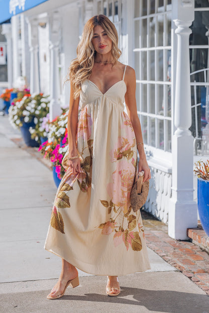 A woman in the Buttercup Floral Print Maxi Dress with clear heels holds a woven clutch on a sunny sidewalk.