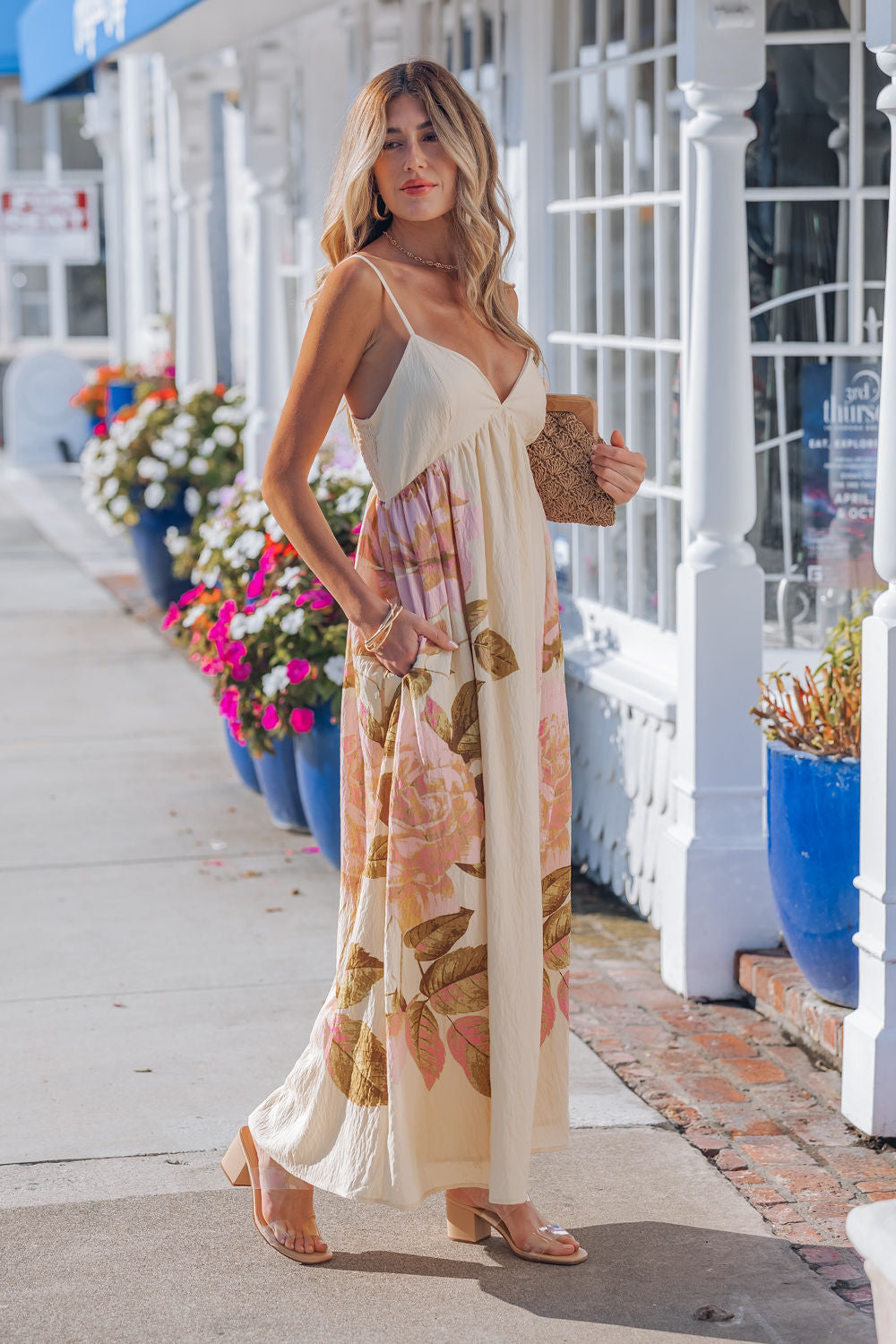 Woman wears the Buttercup Floral Print Maxi Dress with adjustable cami straps by blue flower pots near a white building.