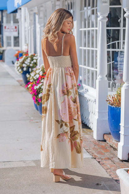 A woman in the Buttercup Floral Print Maxi Dress with adjustable cami straps stands on a sunny sidewalk near flowers and storefronts.