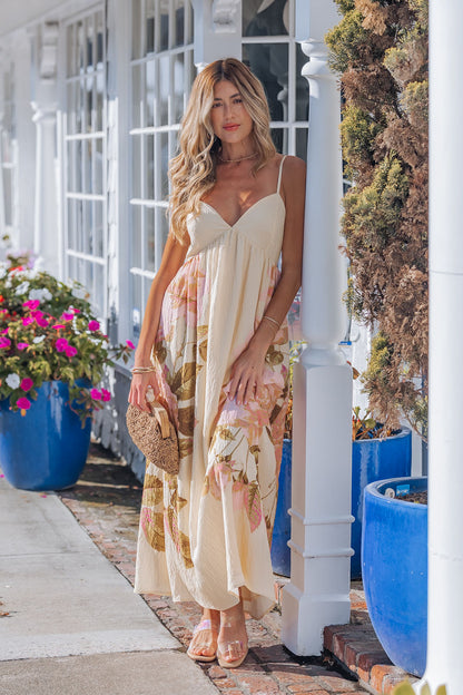 A woman in the Buttercup Floral Print Maxi Dress with adjustable cami straps stands by a white building, holding a woven clutch.