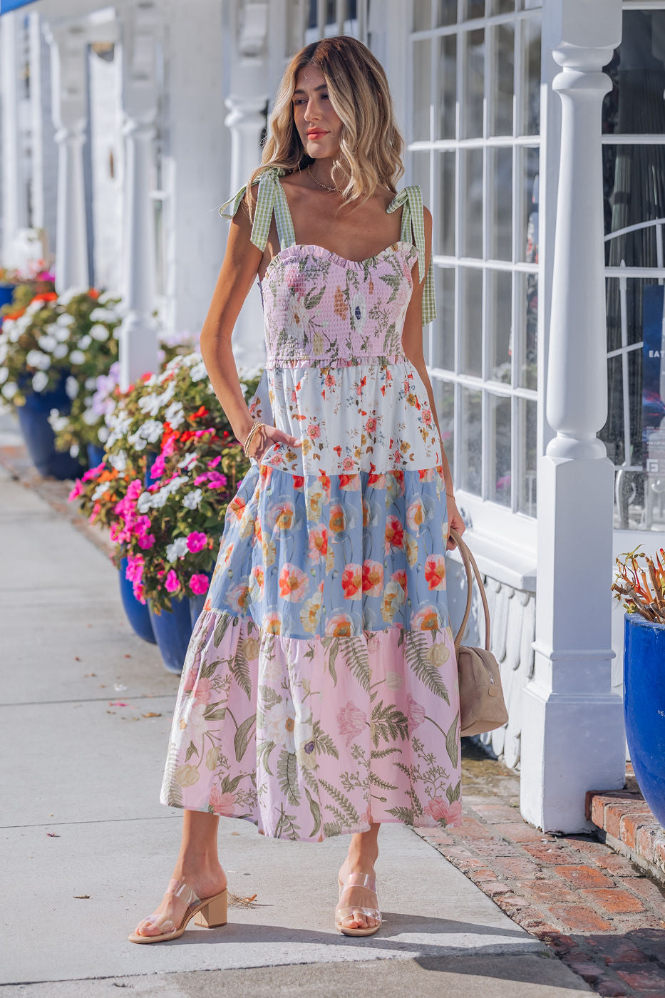 Woman in a Multi Pink Floral Bow Strap Midi Dress stands by blue flower planters, white storefronts behind her.