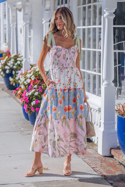 Woman in a Multi Pink Floral Bow Strap Midi Dress stands by blue flower planters, white storefronts behind her.