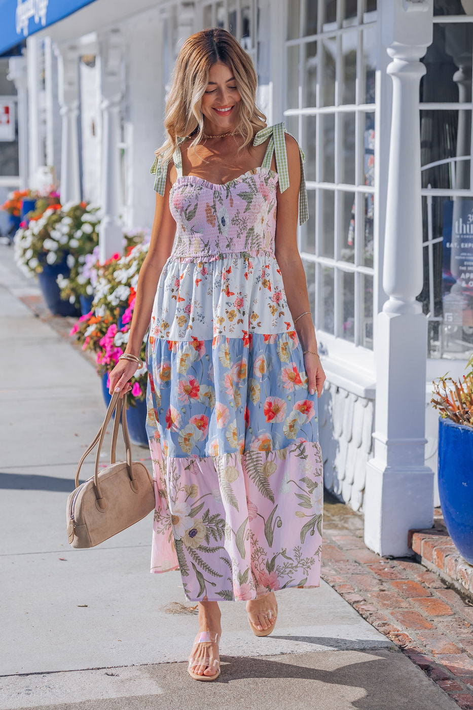 A woman in a Multi Pink Floral Bow Strap Midi Dress walks on a sunny sidewalk, holding a tan bag amid flowers and white buildings.