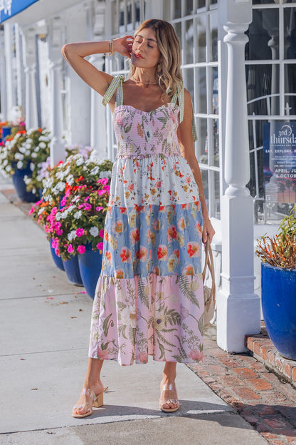 A woman in a Multi Pink Floral Bow Strap Midi Dress and sandals poses on a sunny sidewalk near blue flower pots and white buildings.