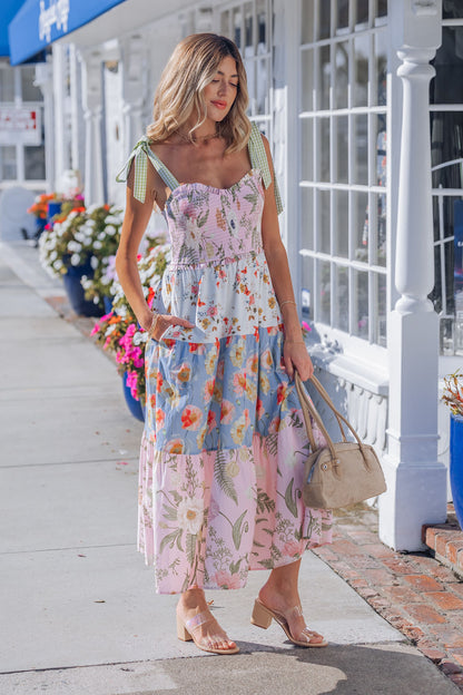 A woman in a Multi Pink Floral Bow Strap Midi Dress walks on a sidewalk, holding a beige bag with colorful potted flowers behind her.