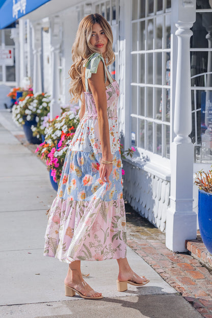 A woman in a Multi Pink Floral Bow Strap Midi Dress and sandals walks past flowers and white storefronts.