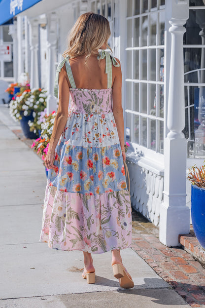 A woman in a Multi Pink Floral Bow Strap Midi Dress strolls down a sunny sidewalk by blooming flowers and white shopfronts.