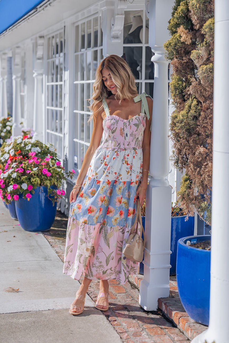 A woman in a Multi Pink Floral Bow Strap Midi Dress leans on a white pillar, with blue flower planters, outdoors on a sunny day.