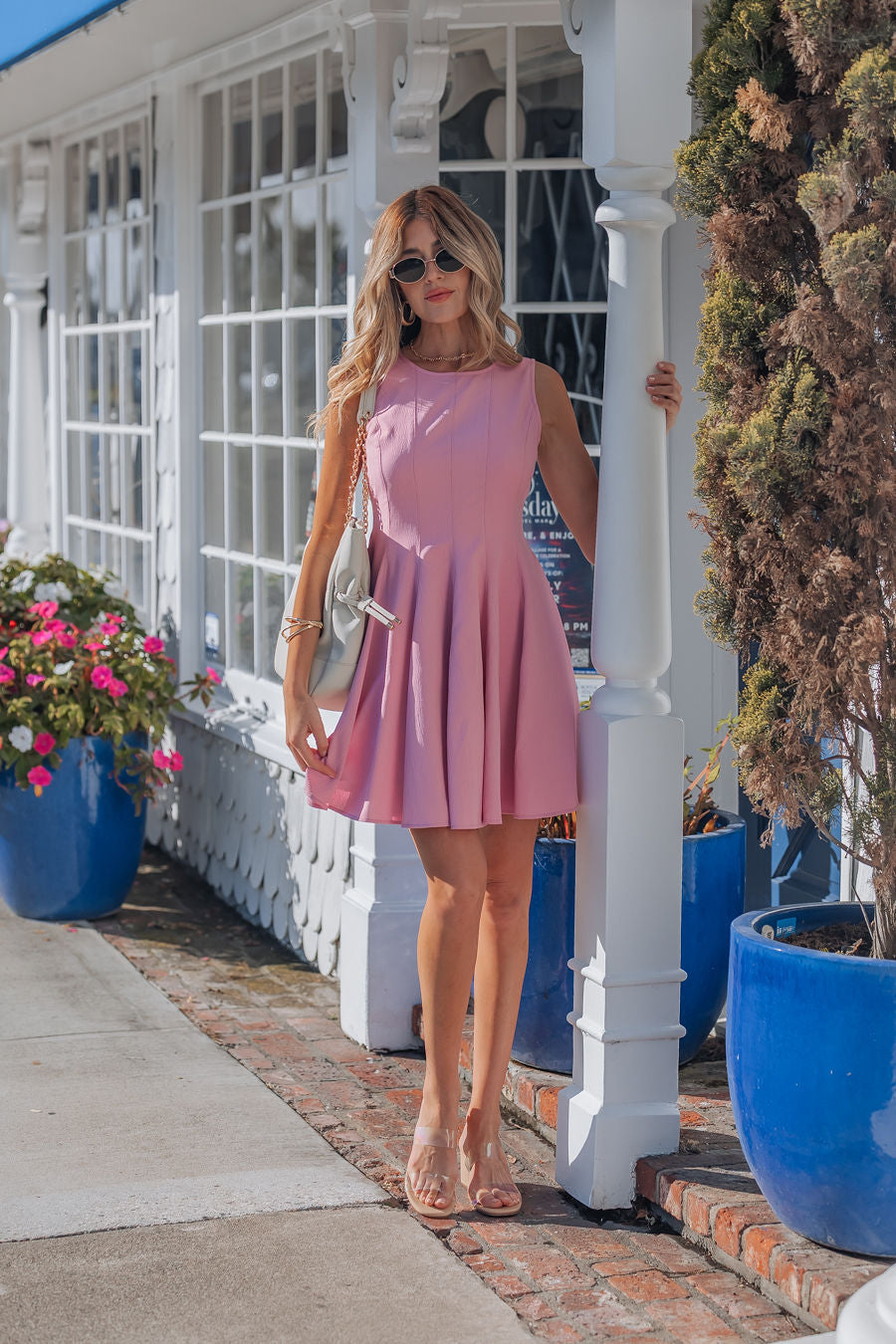 Wearing a Rosette Pink Pleated Mini Dress and sunglasses, a woman stands by a white post near blue flowerpots with blooming plants.