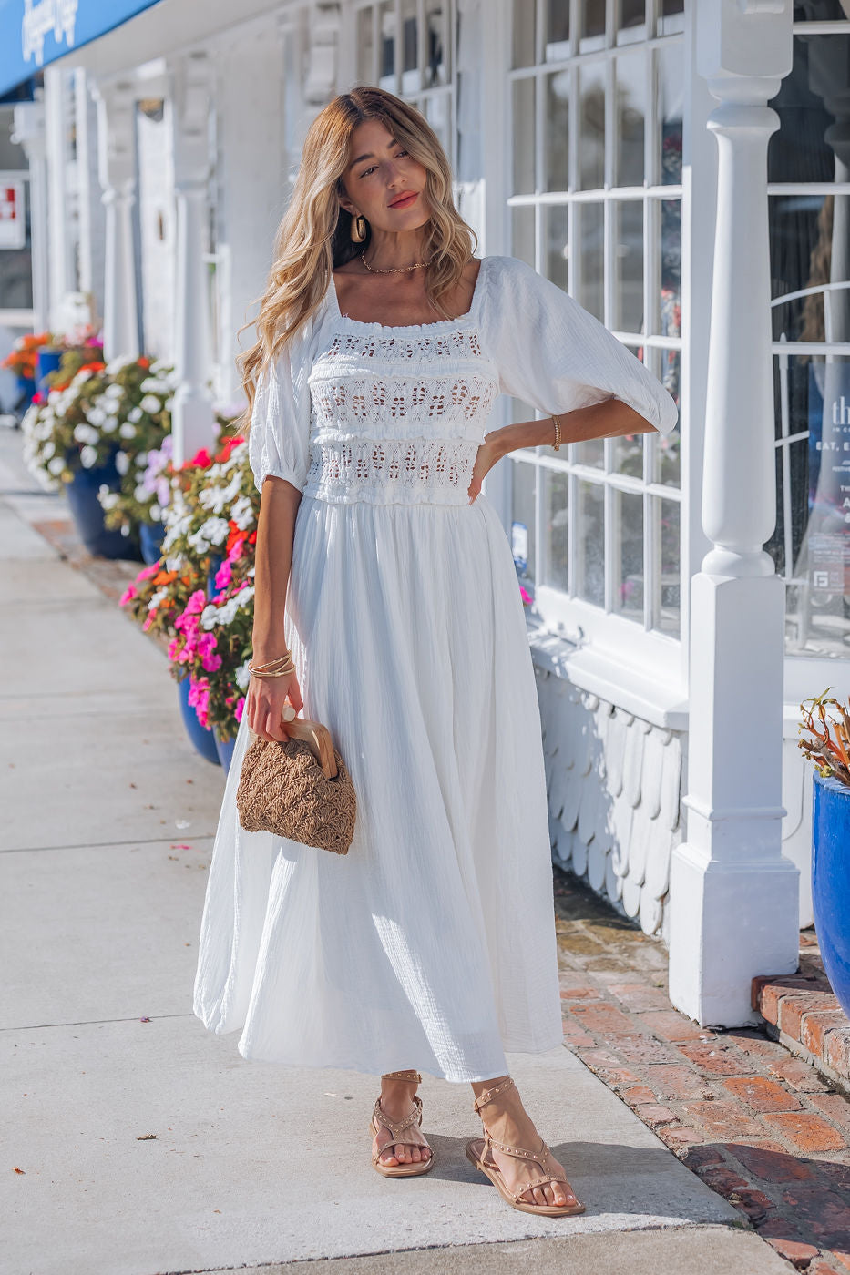 A woman in the Angelica White Ruched Midi Dress holds a woven handbag on a sunny, flower-lined sidewalk.