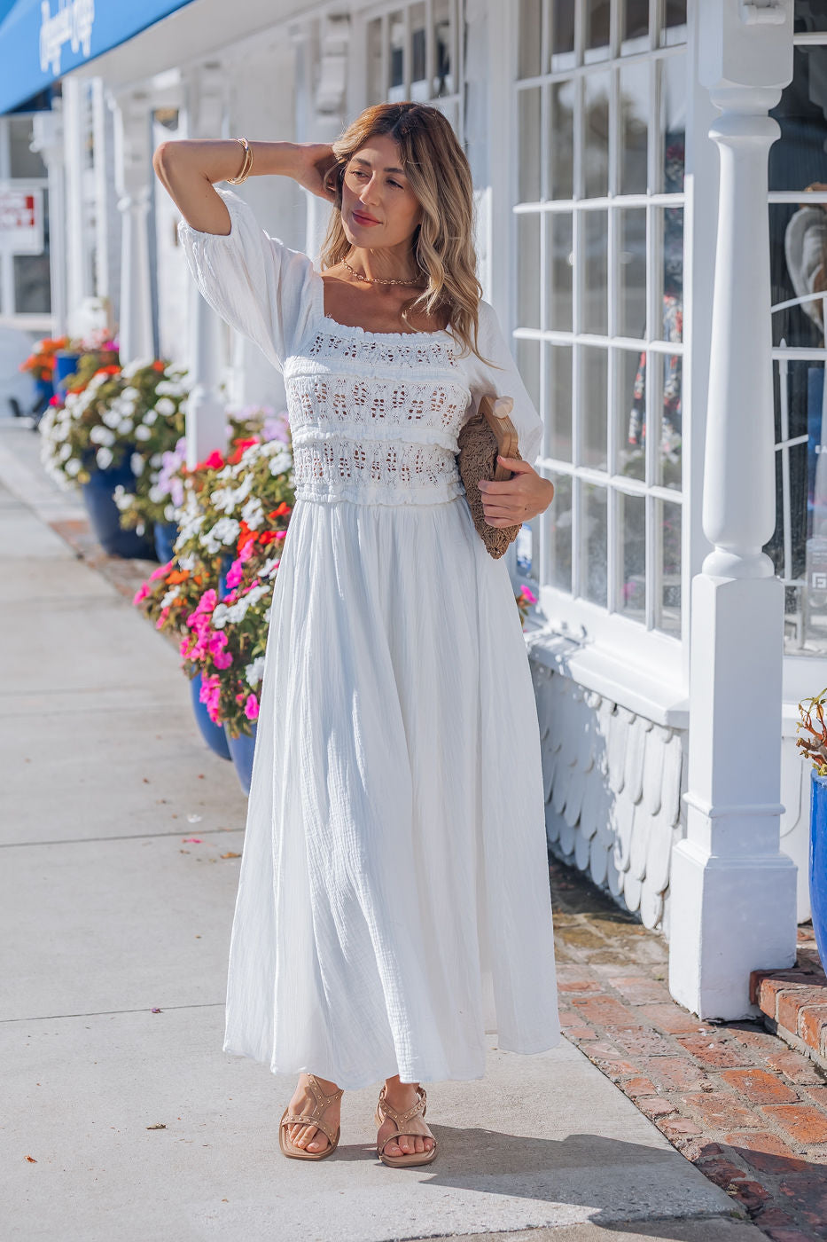 Woman in Angelica White Ruched Midi Dress with puff sleeves and sandals poses on a sunny sidewalk, holding a brown clutch.