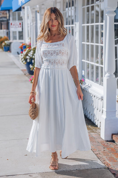 Woman in the Angelica White Ruched Midi Dress with puff sleeves and sandals, carrying a round bag on a sunlit city sidewalk.