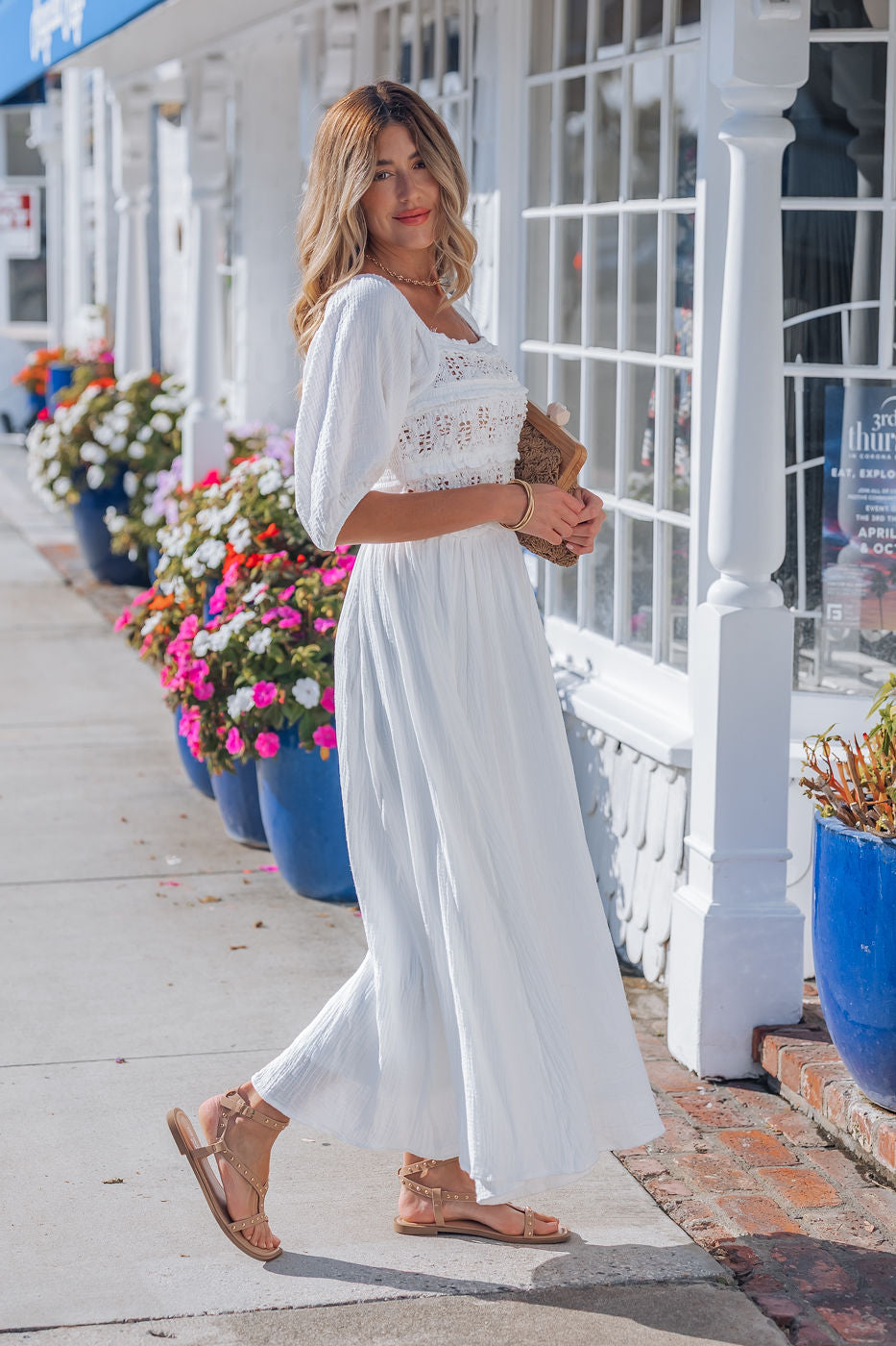 Wearing the Angelica White Ruched Midi Dress, a woman stands by blue pots of flowers, holding a clutch on the sidewalk.