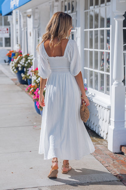 A woman in the Angelica White Ruched Midi Dress and sandals walks on a sunny sidewalk, holding a woven clutch near blue potted flowers.