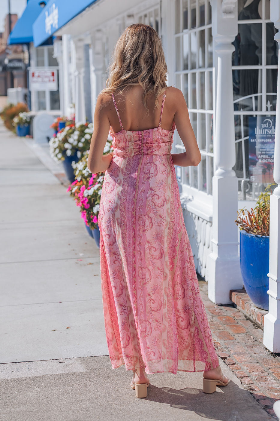 A woman in a Pink Paisley Print Maxi Dress and heels walks on a sunny sidewalk beside a white building with blue planters.