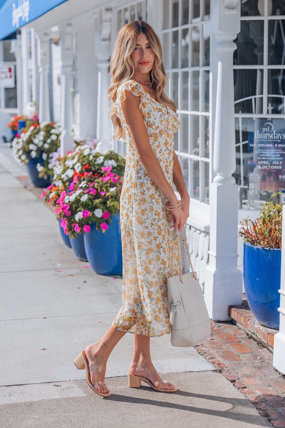 Woman wearing an Ivory Floral Chiffon Midi Dress and nude heels stands on a sidewalk, holding a white handbag.