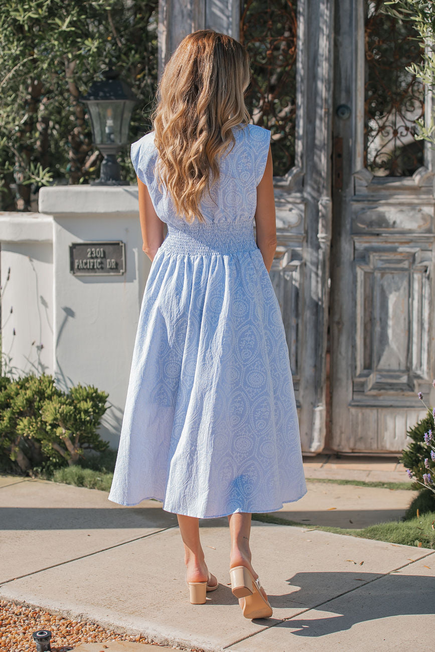 A woman with wavy blonde hair wears a Bluebell Eyelet Smocked Zip Midi Dress and heels, standing outdoors facing away.