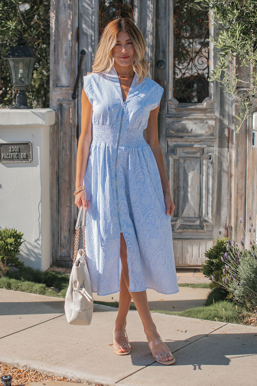 Woman in a Bluebell Eyelet Smocked Zip Midi Dress and sandals holds a white bag by a rustic wooden gate and greenery.