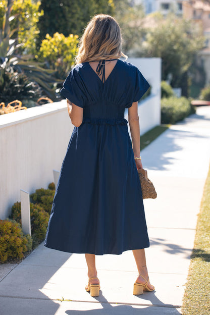 Blonde woman wears a Navy Smocked Zip Midi Dress and yellow sandals on a sunny, greenery-lined sidewalk.