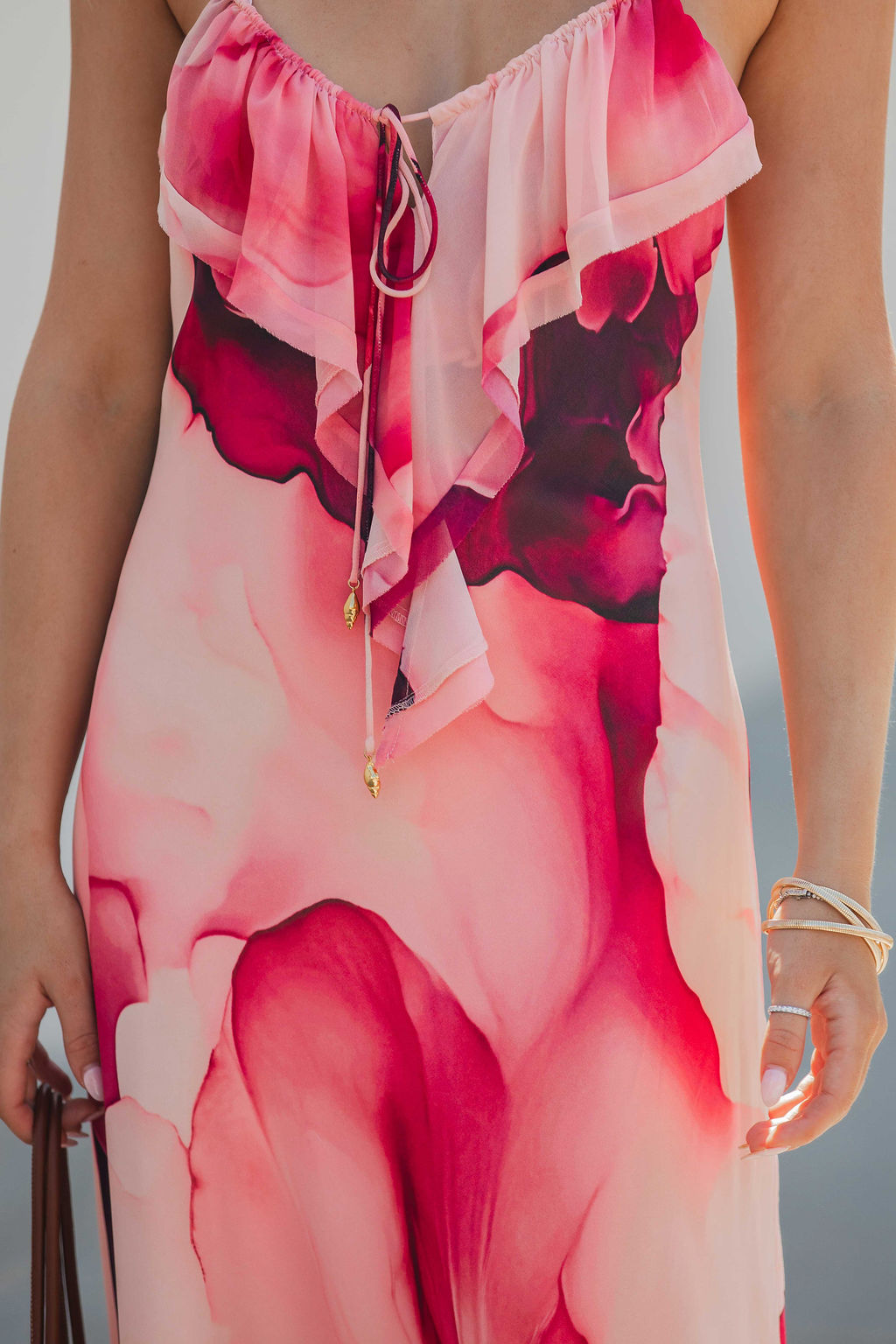 A person wearing the Steve Madden Maxinia Coral Print Maxi Dress with pink-red watercolor patterns, ruffle details, and gold wrist jewelry.