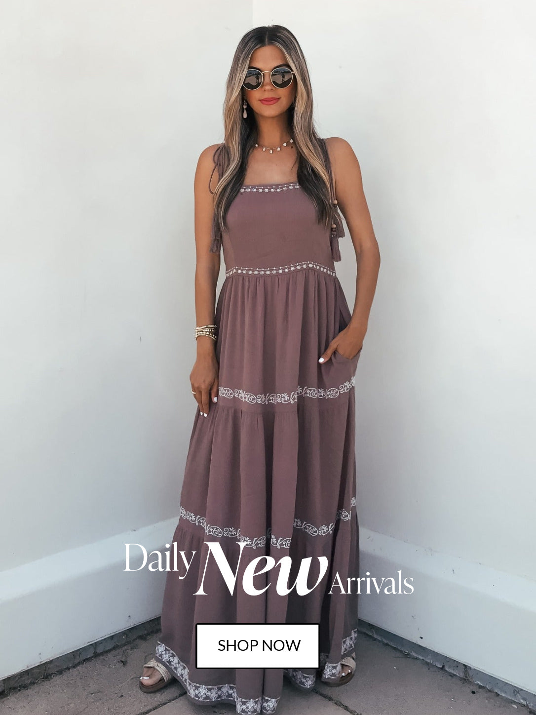 Woman wearing a long brown dress with lace details against a white wall, with text 'Daily New Arrivals' and 'Shop Now'.