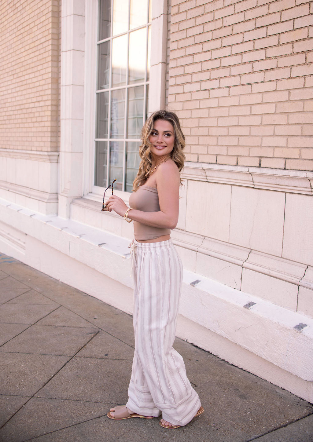 Woman in a beige top and Mocha Striped Palazzo Pants - FINAL SALE stands on a sidewalk holding sunglasses and smiling.