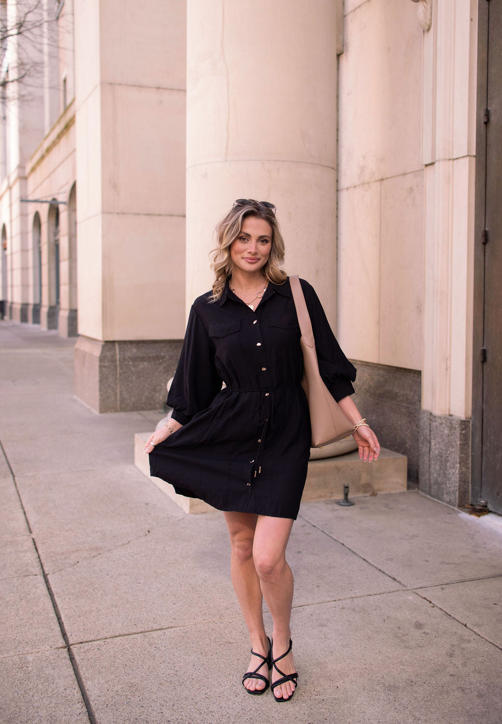 A woman wearing the Black Tie Mini Shirt Dress and sandals stands on a city sidewalk, holding her dress and carrying a beige bag.