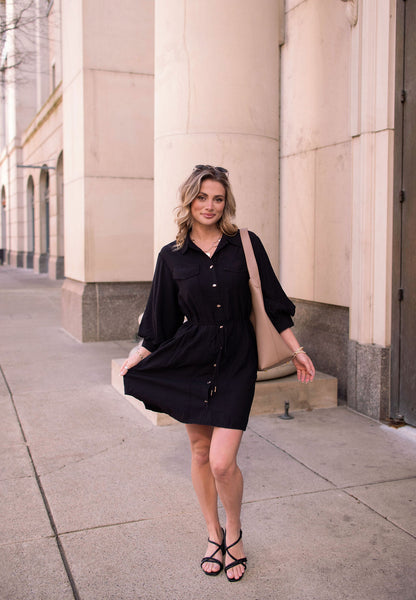 A woman wearing the Black Tie Mini Shirt Dress and sandals stands on a city sidewalk, holding her dress and carrying a beige bag.