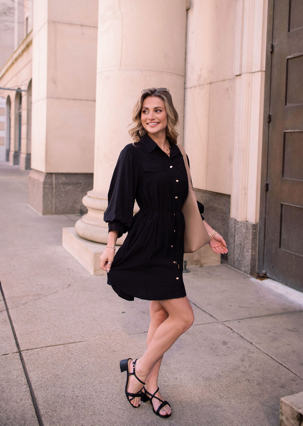 A woman in a Black Tie Mini Shirt Dress and sandals smiles while posing on a city sidewalk near large stone columns.