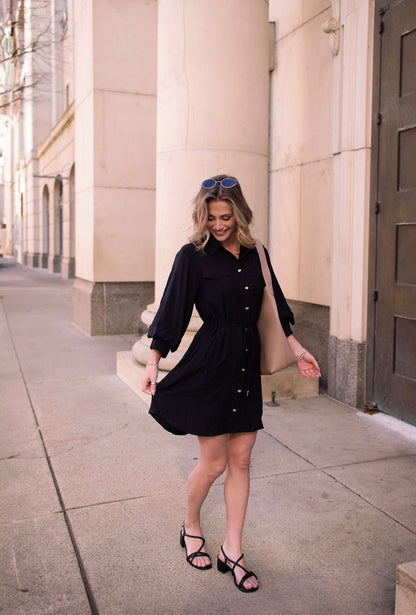 Woman in a Black Tie Mini Shirt Dress and sandals walks city sidewalk, smiling, with sunglasses and a beige tote bag.