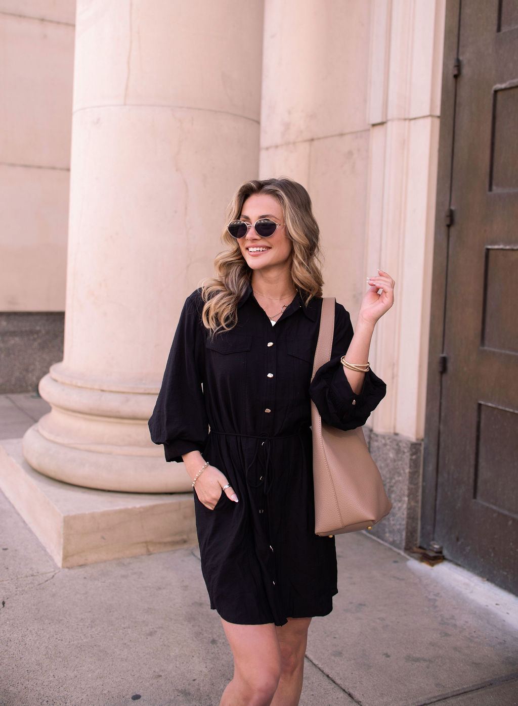 Woman in a Black Tie Mini Shirt Dress, sunglasses, and a beige tote bag smiles before a stone building with columns.