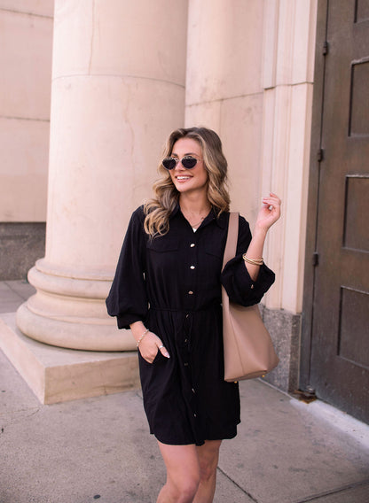 Woman in a Black Tie Mini Shirt Dress, sunglasses, and a beige tote bag smiles before a stone building with columns.
