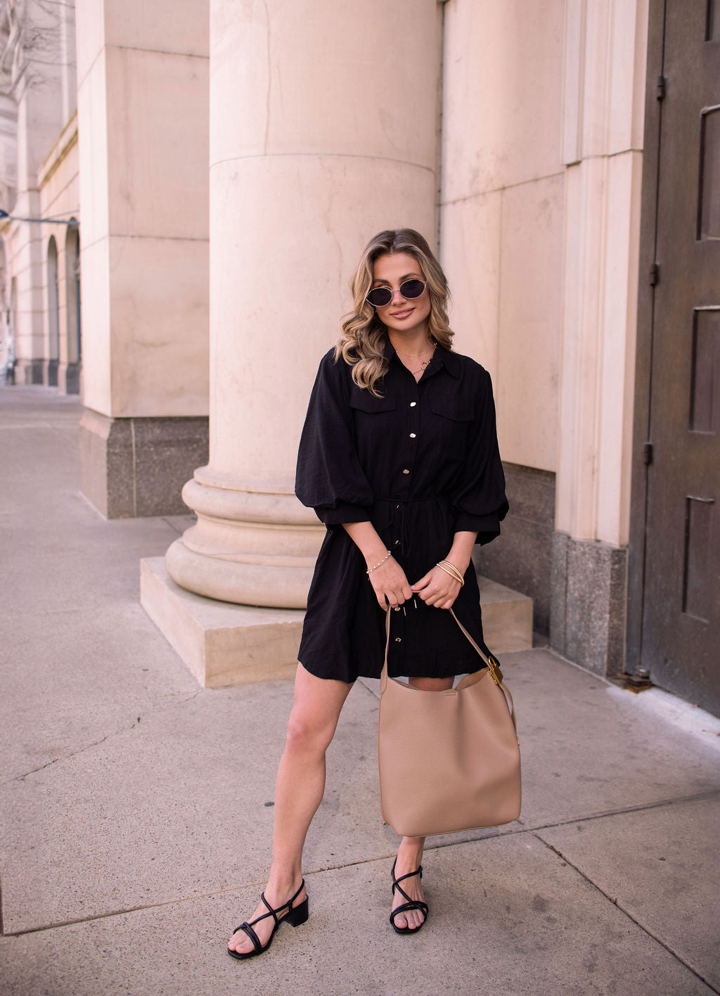 Woman in a Black Tie Mini Shirt Dress, sunglasses, and sandals stands on a sidewalk holding a tan handbag near tall columns.