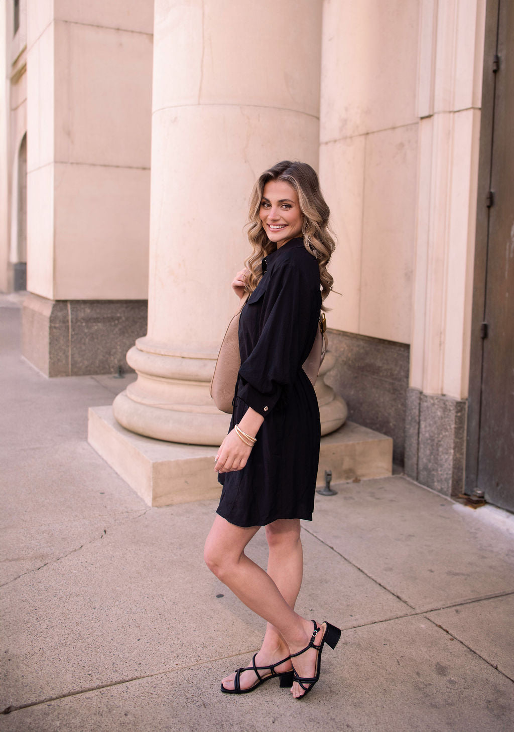 A woman in the Black Tie Mini Shirt Dress and sandals smiles near stone columns, holding a bag over her shoulder.