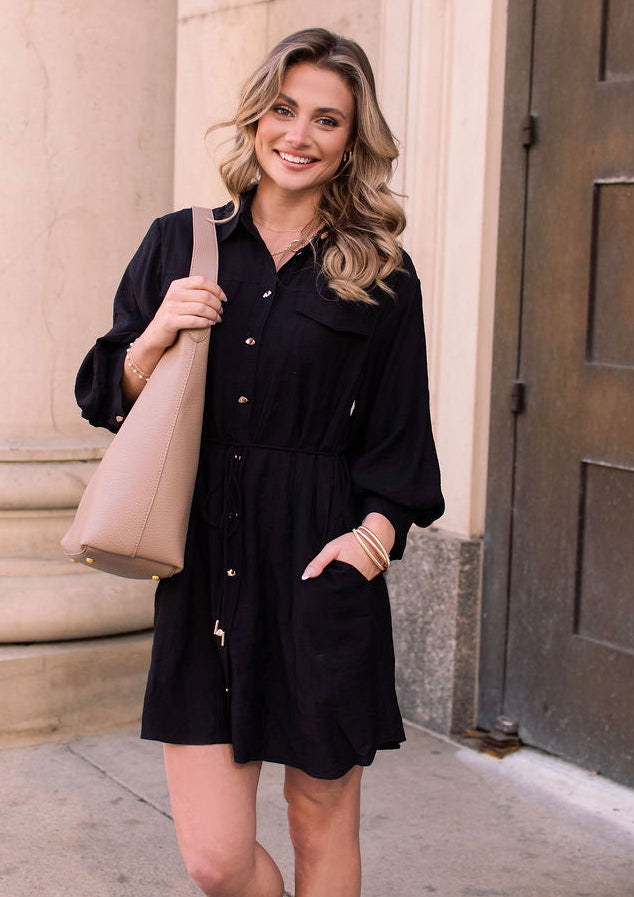 A woman with wavy hair wears the Black Tie Mini Shirt Dress and sandals, holding a beige tote by a stone column and large door.