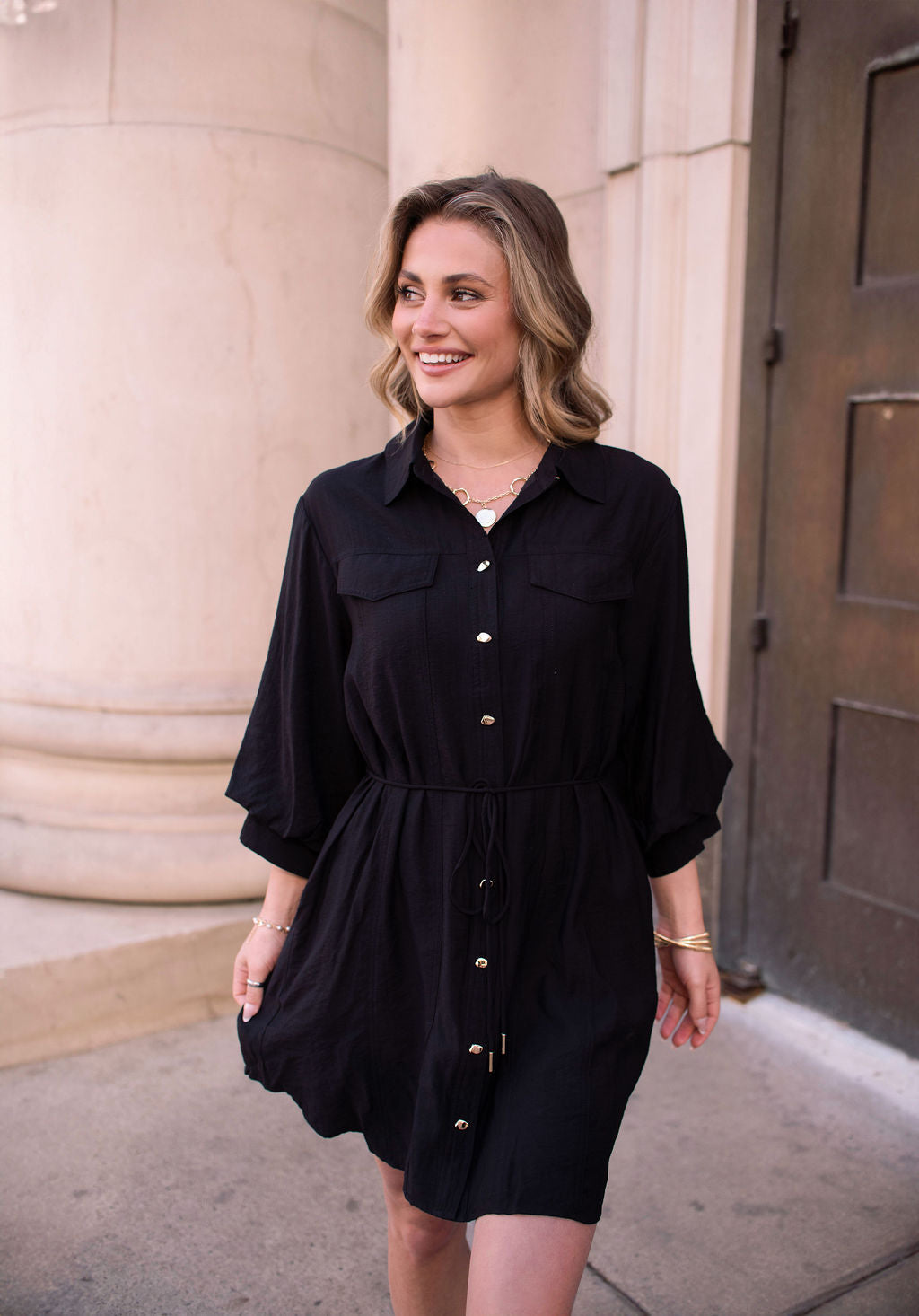 A woman in the Black Tie Mini Shirt Dress smiles as she walks outside near a column and a dark door.
