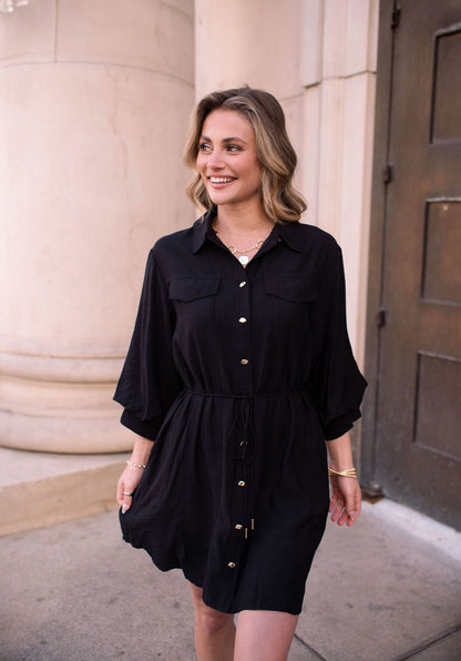 A woman in the Black Tie Mini Shirt Dress smiles as she walks outside near a column and a dark door.