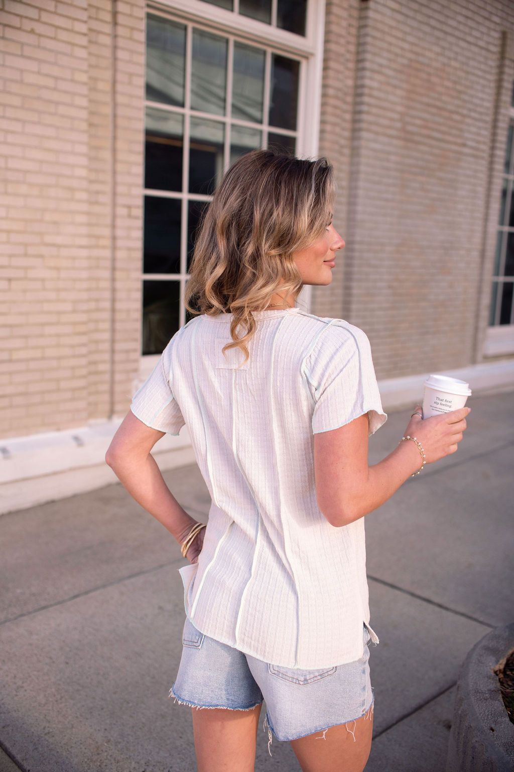 A woman with wavy hair stands outdoors in the Natural Contrast Trim Button Up Top, holding a coffee cup near a brick building.