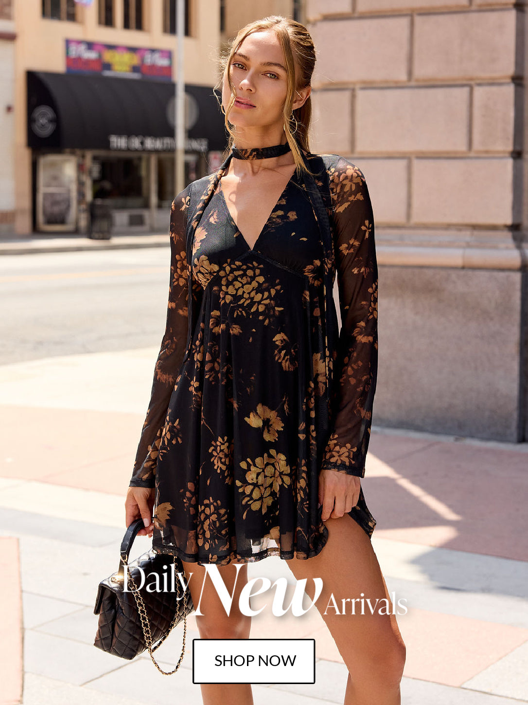 A woman modeling a black long-sleeve mini dress with a gold floral pattern, standing on a city sidewalk. The dress features a V-neckline, sheer sleeves, and a flared skirt.