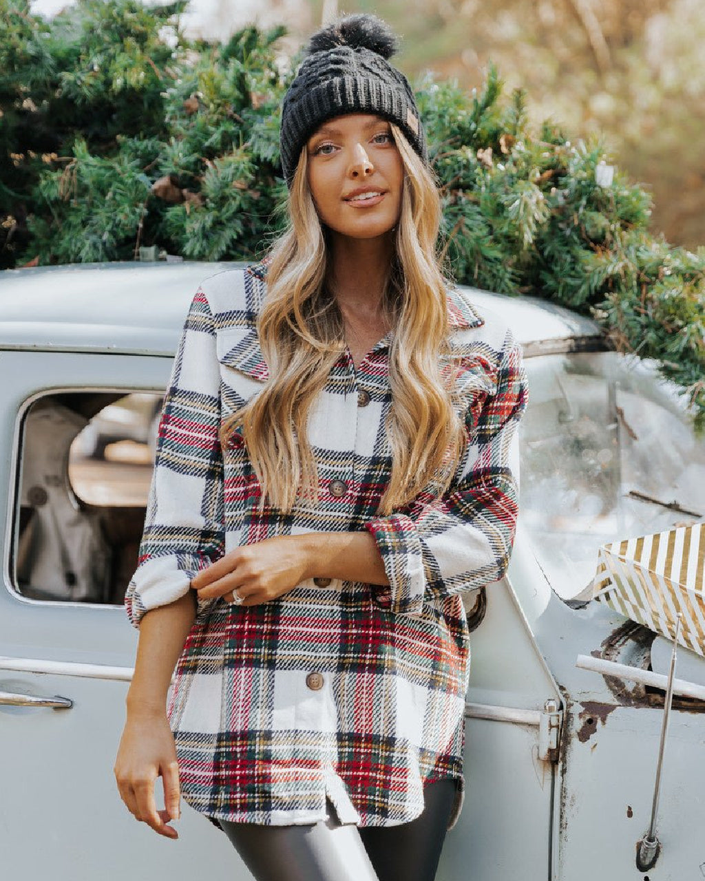 Woman wearing a plaid shirt and beanie standing next to a vintage van with greenery in the background