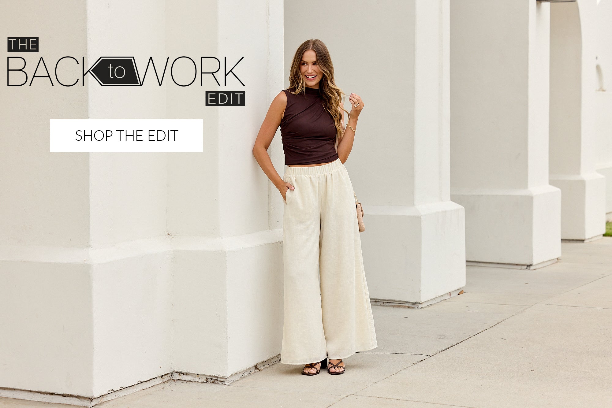 A woman stands outdoors against a white architectural wall wearing a sleeveless dark plum top and wide-leg cream pants. Text in the upper left reads 'THE BACK to WORK EDIT' with a 'SHOP THE EDIT' button below it.