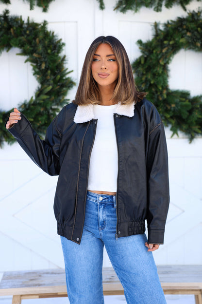A woman in a Black Shearling Collar Faux Leather Jacket and blue jeans stands before a white wall with green wreaths.