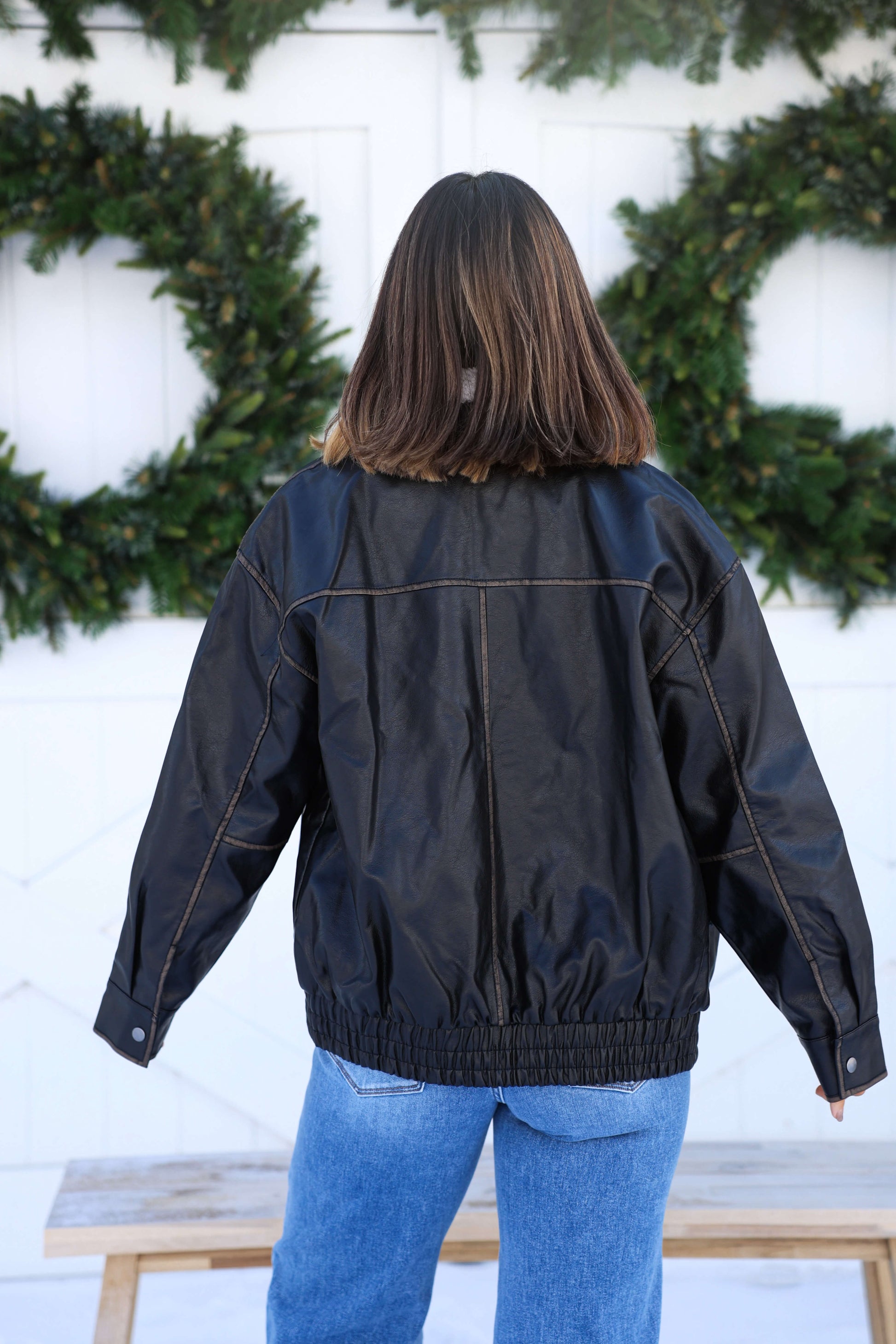 A person with shoulder-length hair wears the Black Shearling Collar Faux Leather Jacket and blue jeans, facing away by a white wall with greenery.