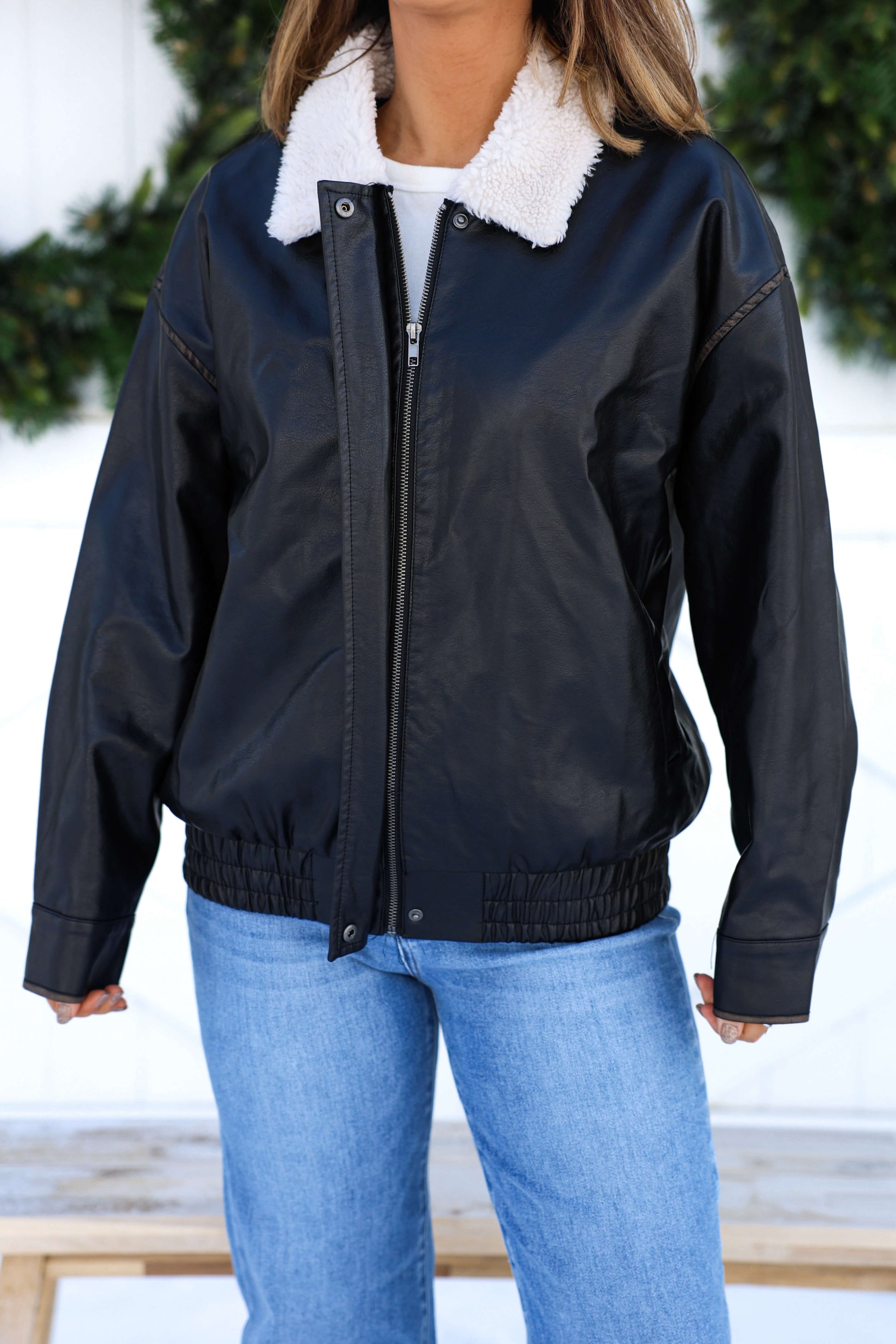 A woman wears a Black Shearling Collar Faux Leather Jacket with blue jeans, standing before greenery and exuding casual cool style.